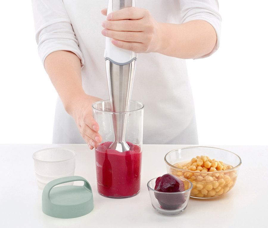Frau püriert Smoothie im Mixbecher. Daneben stehen eine To-go-Lunchbox und Schüsseln mit Zutaten.