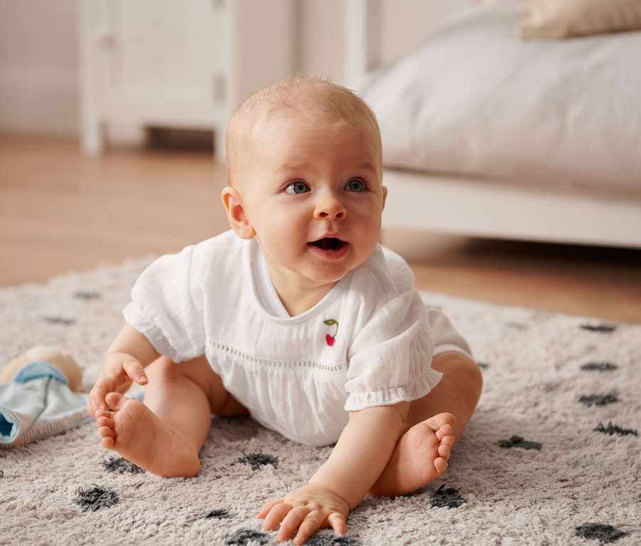 Baby sitzt auf einem Teppich und trägt eine Bluse.