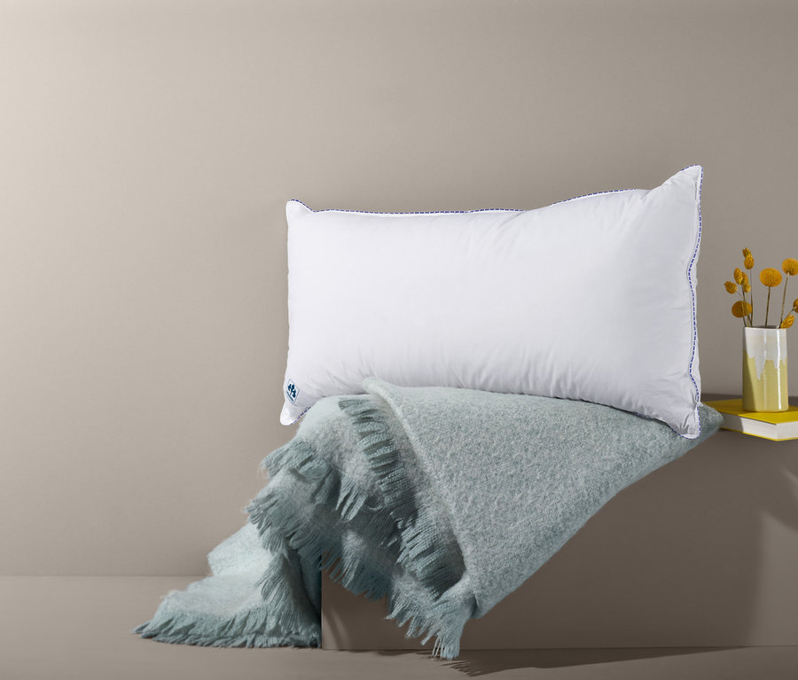Ein weißes irisette® Visco-3-Kammerkissen mit Daunen und Federn liegt auf einer hellblauen, zotteligen Decke. Daneben liegen ein gelbes Buch und eine Vase mit gelben Blumen.