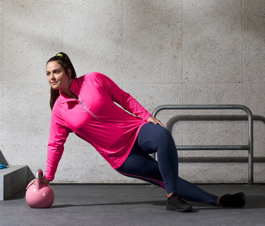 Frau macht Übungen mit einer Kettlebell, sie trägt eine Sporttight und ein neonpinkes Thermo-Funktionsshirt.