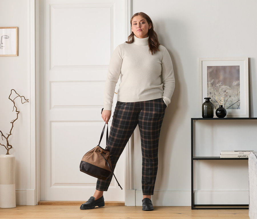 Frau posiert in Cashmere-Feinstrickpullover, karierten Joggpants, Leder-Loafer und Umhängetasche.