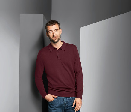 Mann lehnt an Wand und trägt einen bordeauxroten Merino-Pullover.
