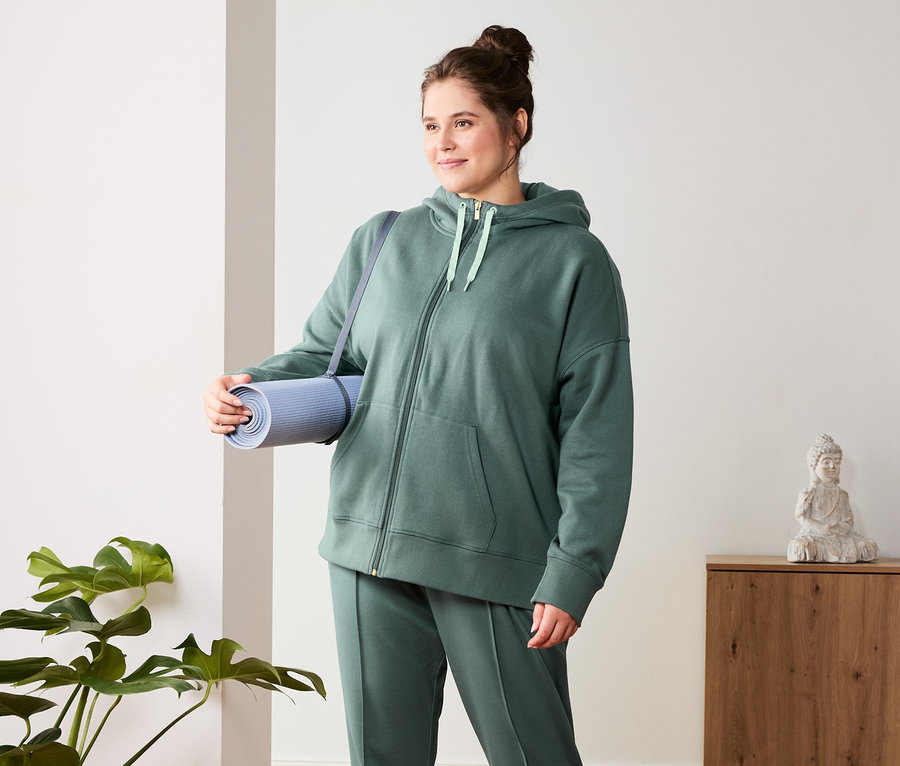 Frau hält Yogamatte und trägt eine dunkelgrüne Sweathose und eine Sweatjacke.