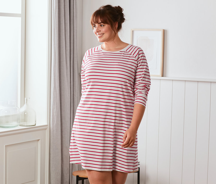 Frau steht am Fenster und trägt ein cremefarben-rot gestreiftes Nachthemd.