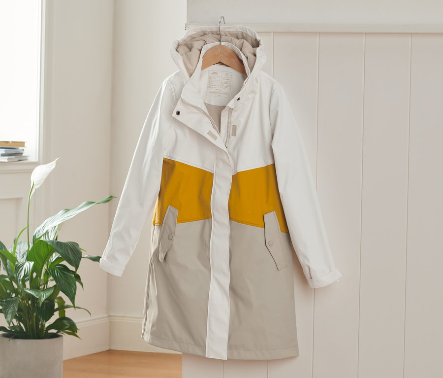 Ein weißer, gelber und beiger Regenparka hängt auf einem Kleiderbügel an einer weißen Wand.