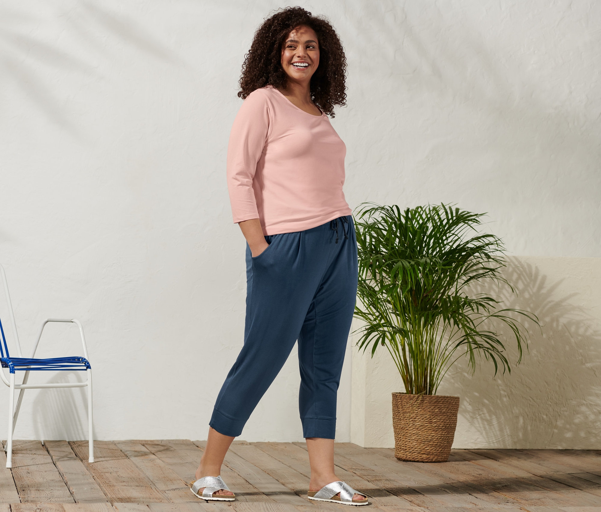 Lächelnde Frau posiert in roséfarbenem Shirt mit 3/4-Arm, 3/4-Jerseyhose und Komfortsandale.