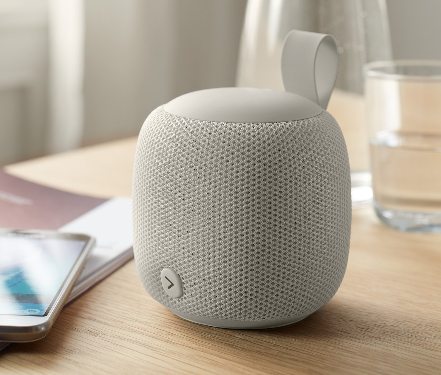 Ein kleiner, grauer Fabric-Bluetooth®-Lautsprecher steht neben einem Handy auf einem Tisch.