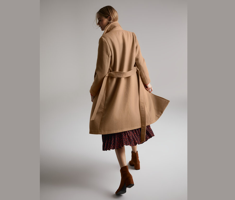 Frau geht in einem camel Mantel aus weicher Wollmischung, rotem Kleid und braunen Stiefeln.