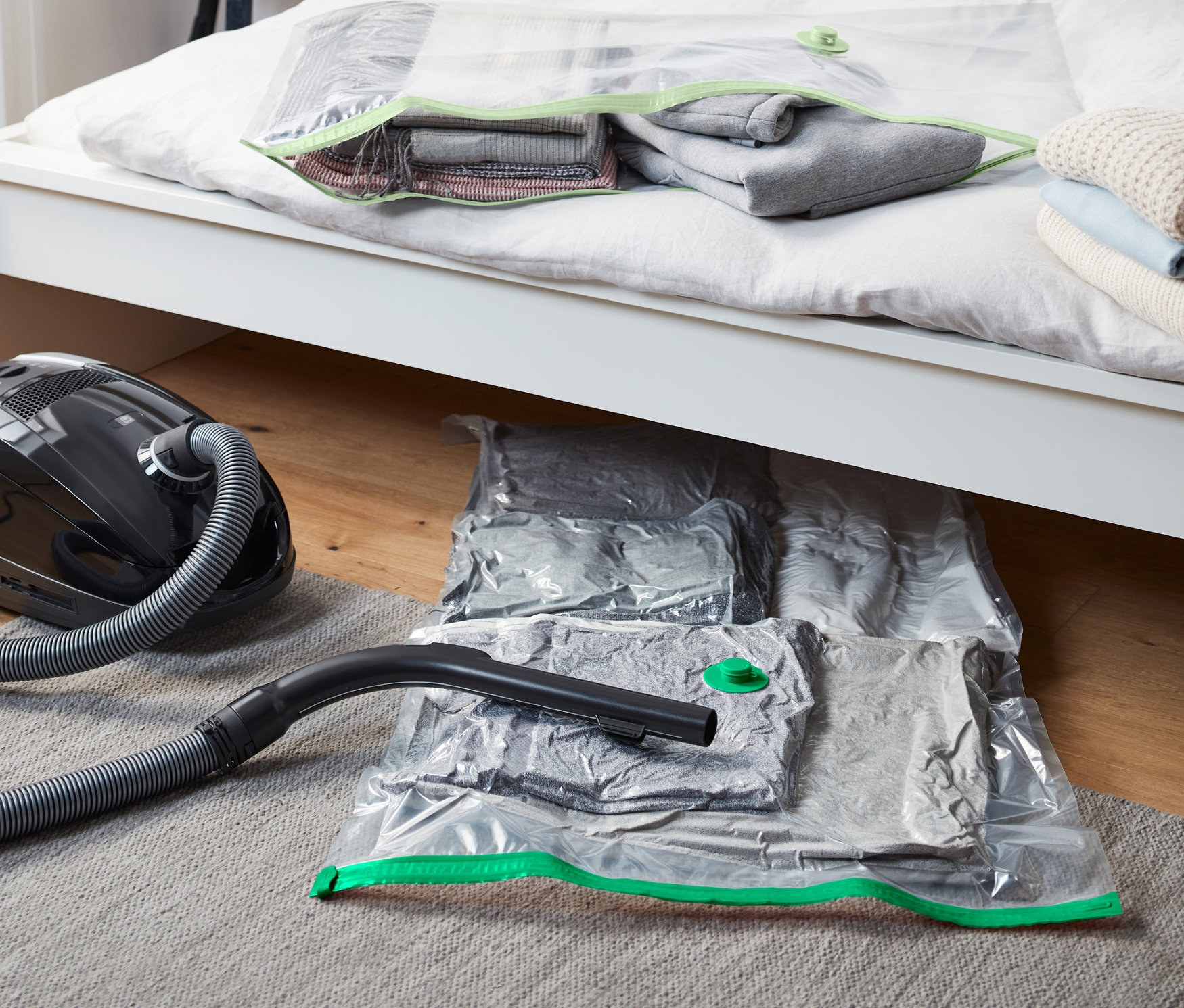 Schwarzer Staubsauger steht neben Bett. Auf und unter dem Bett liegen 2 Vakuum-Aufbewahrungsbeutel, liegend.