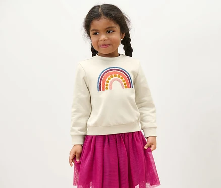 Mädchen trägt einen Kinder-Sweater mit Regenbogen-Applikation und einen lila Rock.
