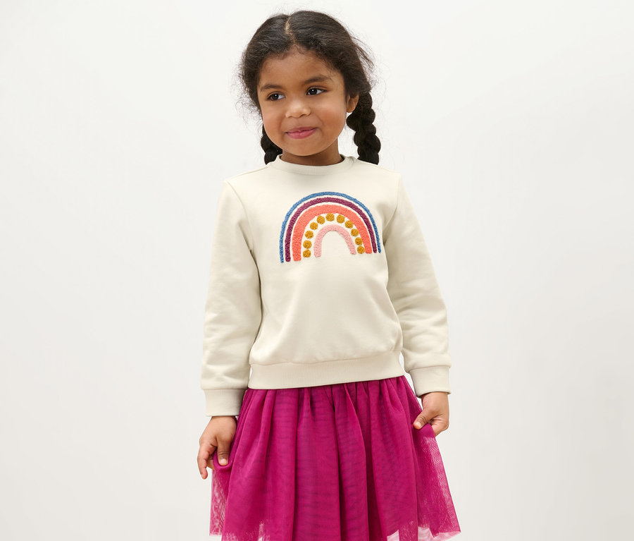 Mädchen trägt einen Kinder-Sweater mit Regenbogen-Applikation und einen lila Rock.