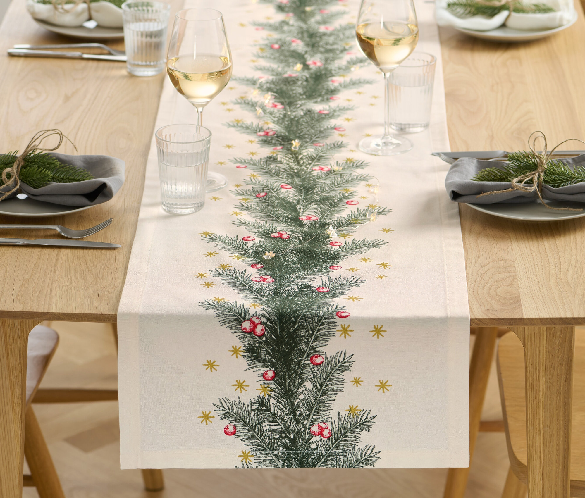 Ein Tischläufer mit weihnachtlichem Print liegt in der Mitte eines Holztisches. Auf beiden Seiten befinden sich Gedecke mit Tellern, Gläsern und Besteck.