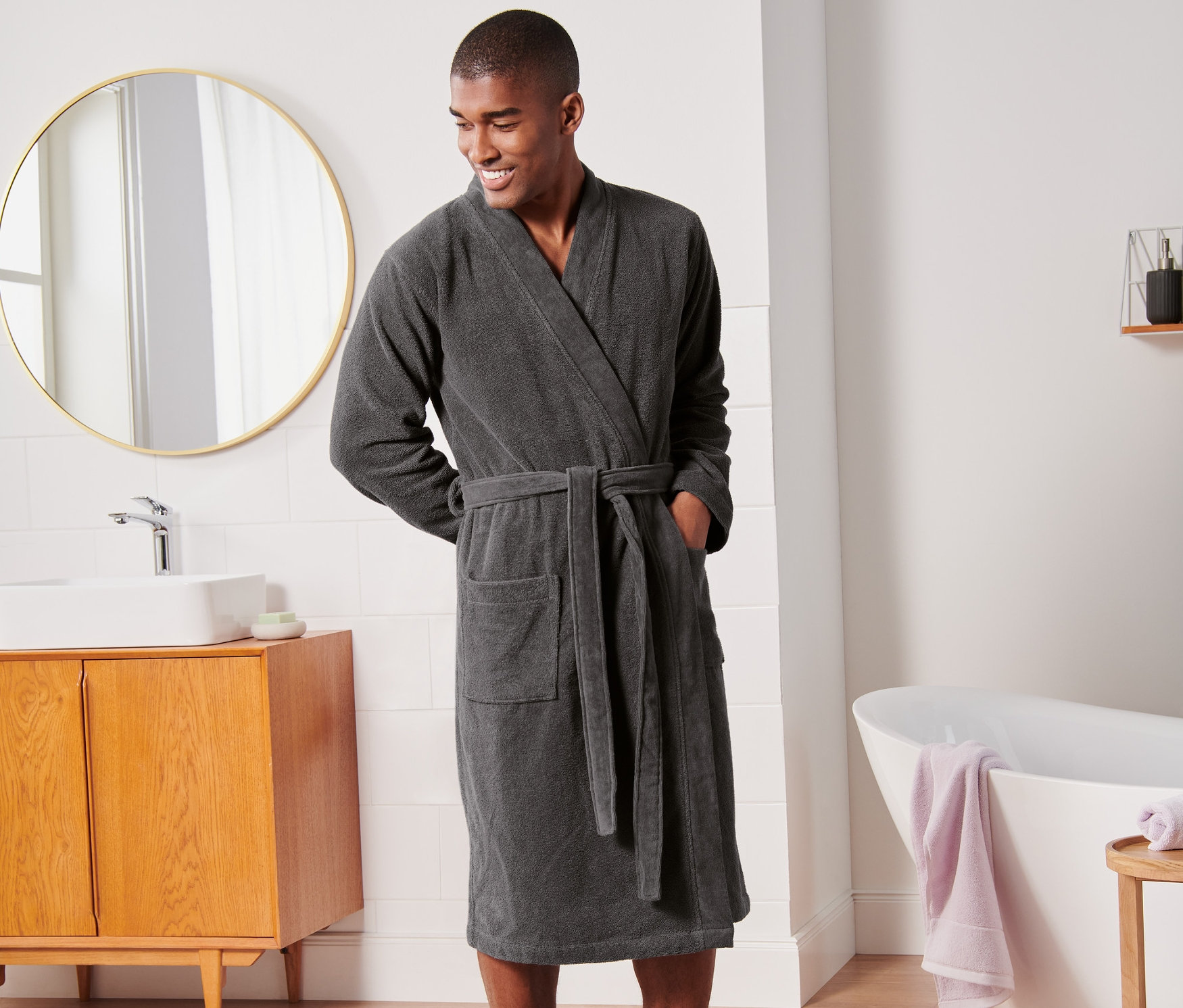 Ein lächelnder Mann steht im Badezimmer und trägt einen anthrazitfarbenen Premium-Bademantel.