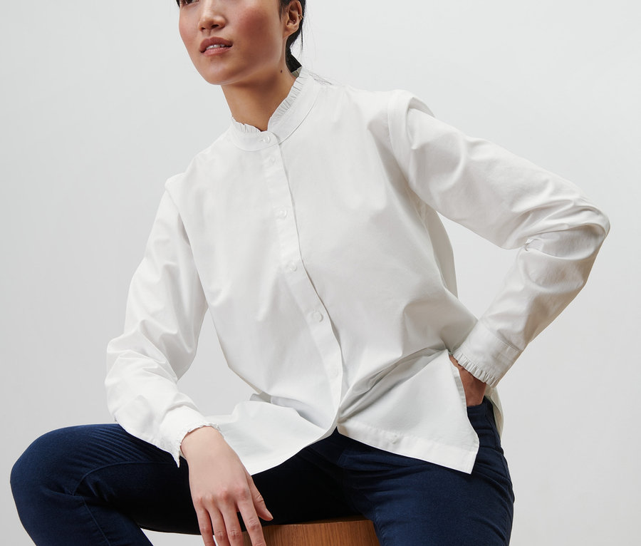 Asiatische Frau sitzt auf einem Holzhocker und trägt eine weiße Bluse mit Rüschendetail und dunkelblaue Hosen.