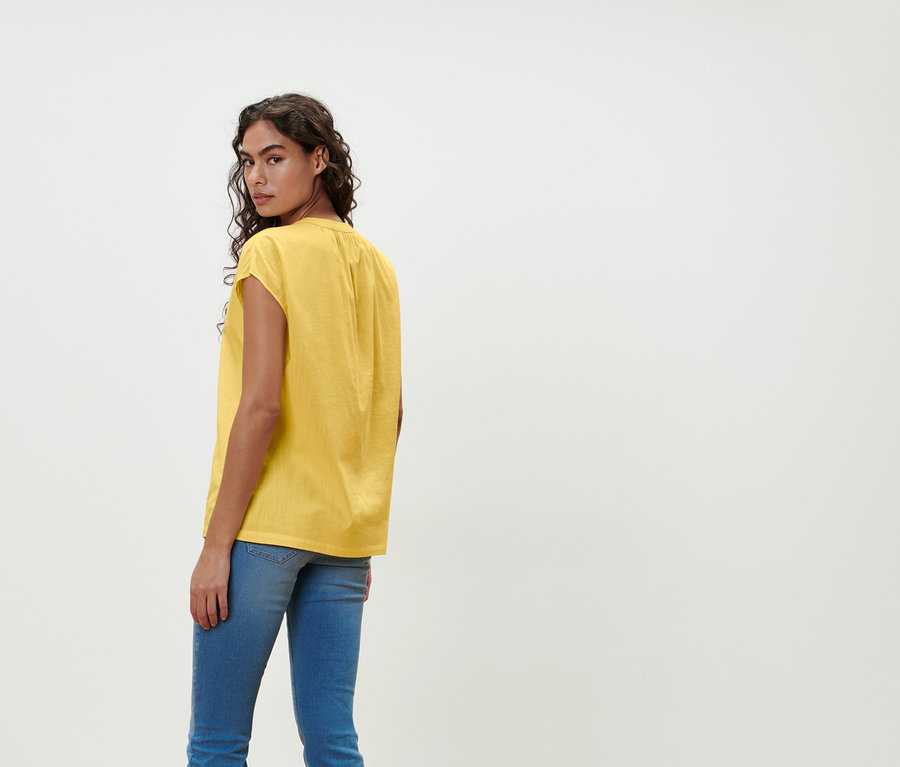 Eine Frau blickt über die Schulter und trägt eine gelbe Kurzarmbluse und Jeans.