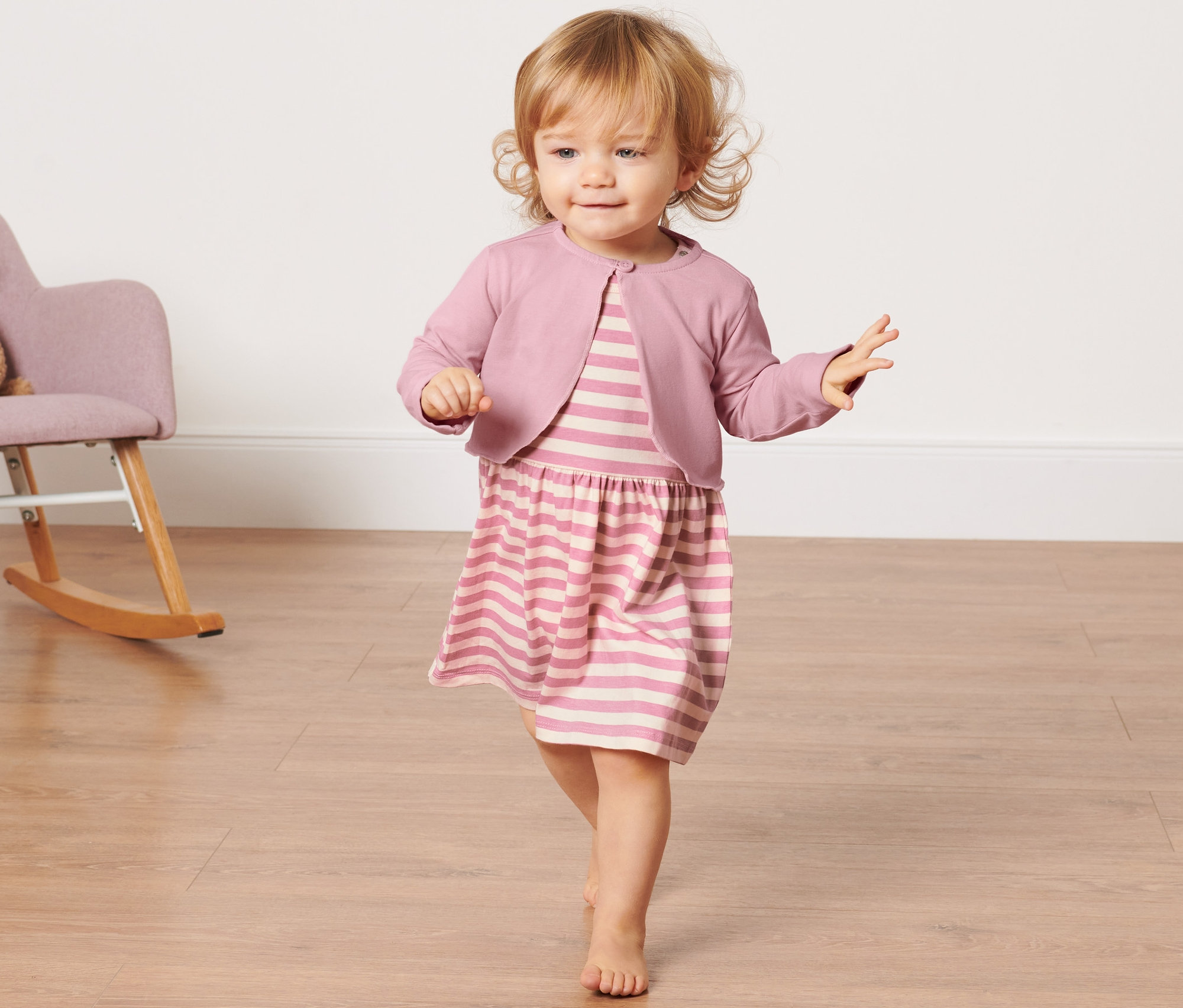 Kleinkind in gestreiftem Kleid und Baby-Bolero-Jacke geht auf Holzboden. Im Hintergrund steht ein Schaukelstuhl mit Teddybär.