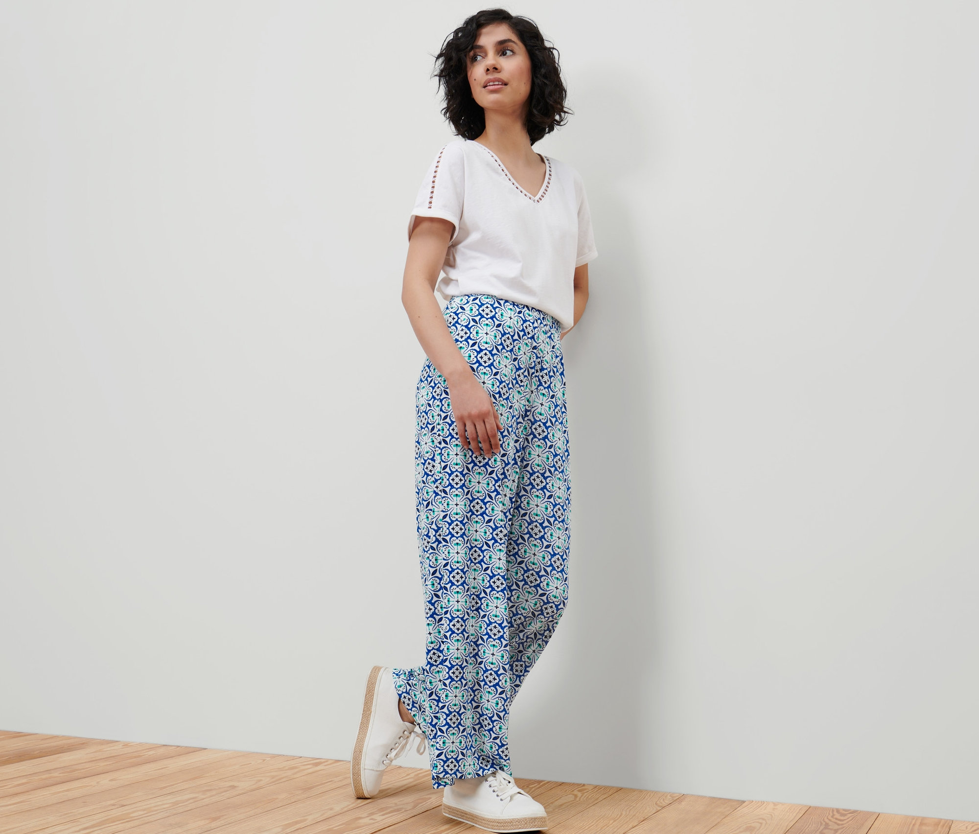 Eine Frau steht an einer Wand und trägt eine weiße Bluse und eine Palazzohose mit Alloverprint.