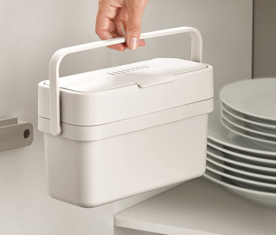 Eine Hand hält einen weißen Joseph Joseph Küchenkomposter neben einer Schranktür.