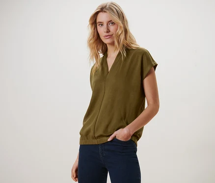 Blonde Frau posiert in einem olivgrünen Blusenshirt mit V-Ausschnitt und dunkelblauen Jeans.