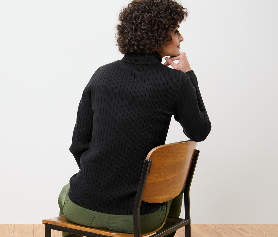 Frau sitzt auf einem Stuhl und trägt einen gerippten schwarzen Rollkragenpullover.