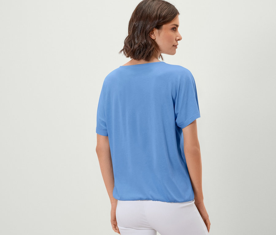 Frau steht mit dem Rücken zur Kamera und trägt ein hellblaues Blusenshirt.