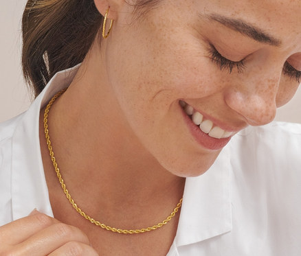 Eine lächelnde Frau schaut nach unten und berührt eine goldene Kordel Kette aus 925er Silber vergoldet. Sie trägt Gold-Creolen Basic flexibel und einen goldenen V-Ring.