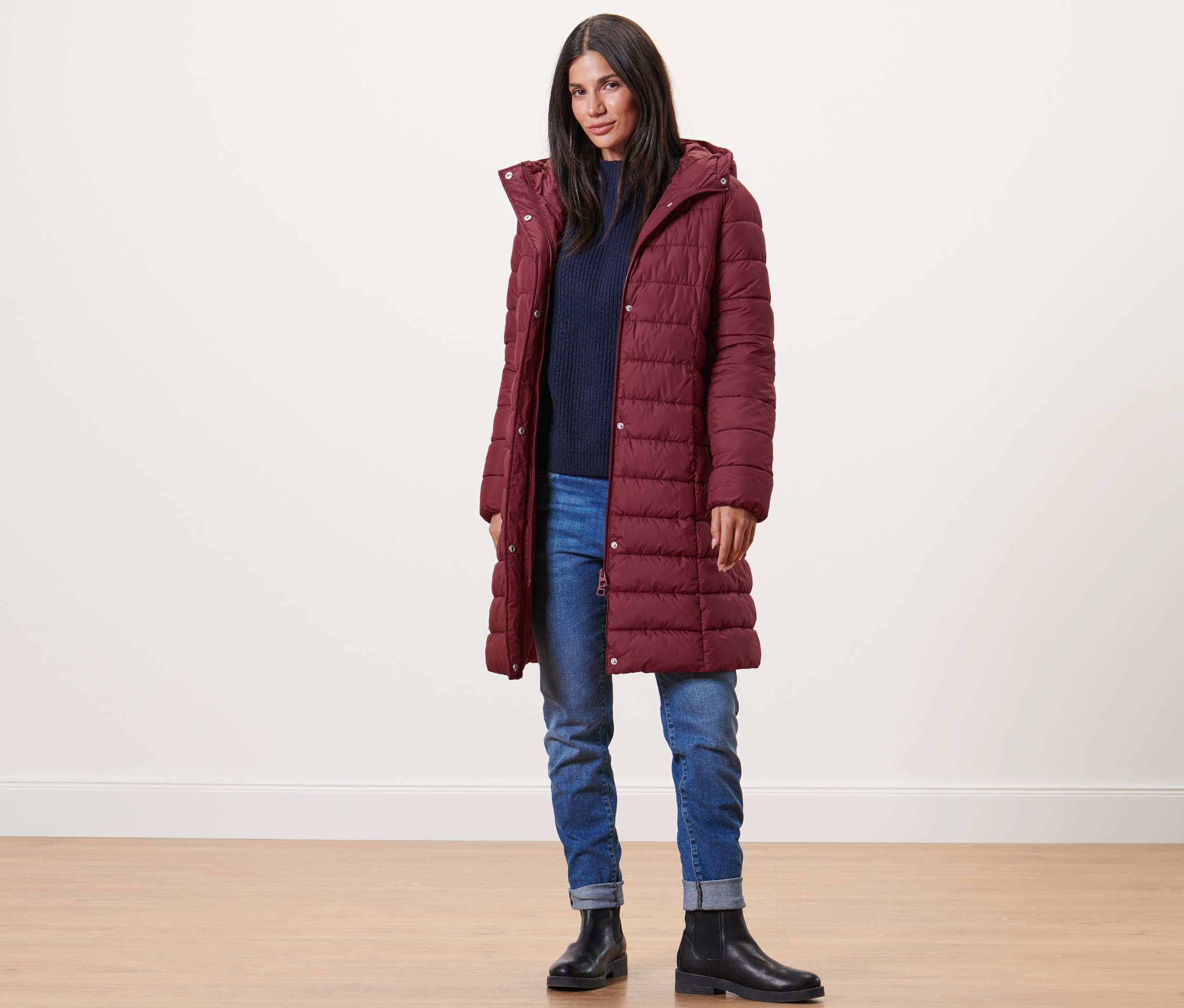 Frau trägt einen bordeauxroten Steppmantel mit Kapuze, einen blauen Pullover, Jeans und schwarze Stiefel.