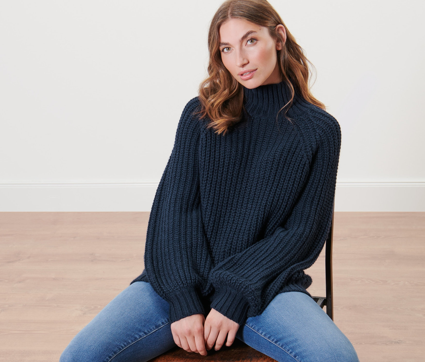 Frau sitzt auf einem Stuhl und trägt einen dunkelblauen Strickpullover mit Stehkragen.