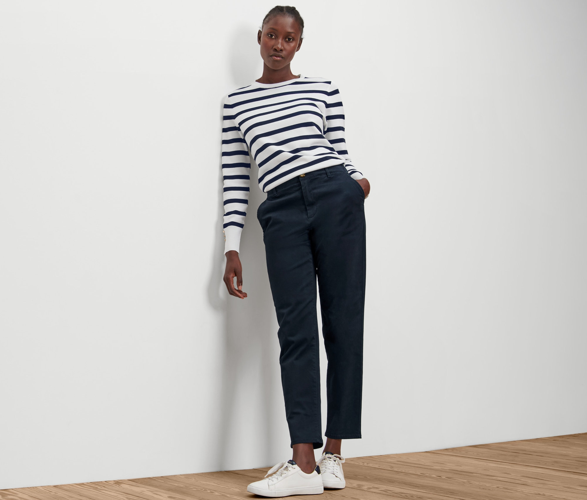 Eine Frau lehnt an einer Wand und trägt ein gestreiftes Oberteil und eine marineblaue Chino.