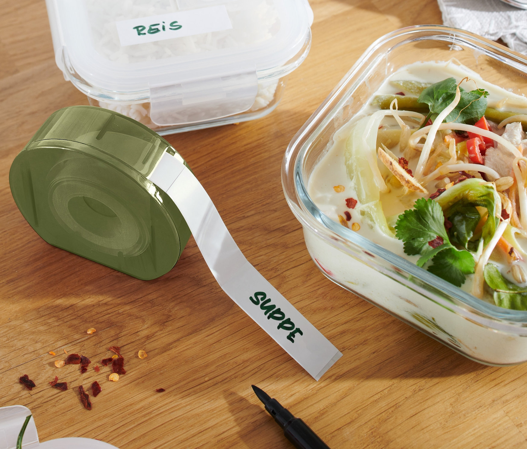 Ein Etikettenspender mit der Aufschrift "SUPPE" liegt neben einem Glasbehälter mit Suppe und einem weiteren Behälter mit Reis.