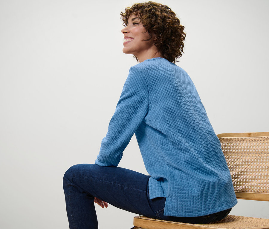 Frau sitzt auf einem Stuhl und trägt ein blaues, strukturiertes Sweatshirt.