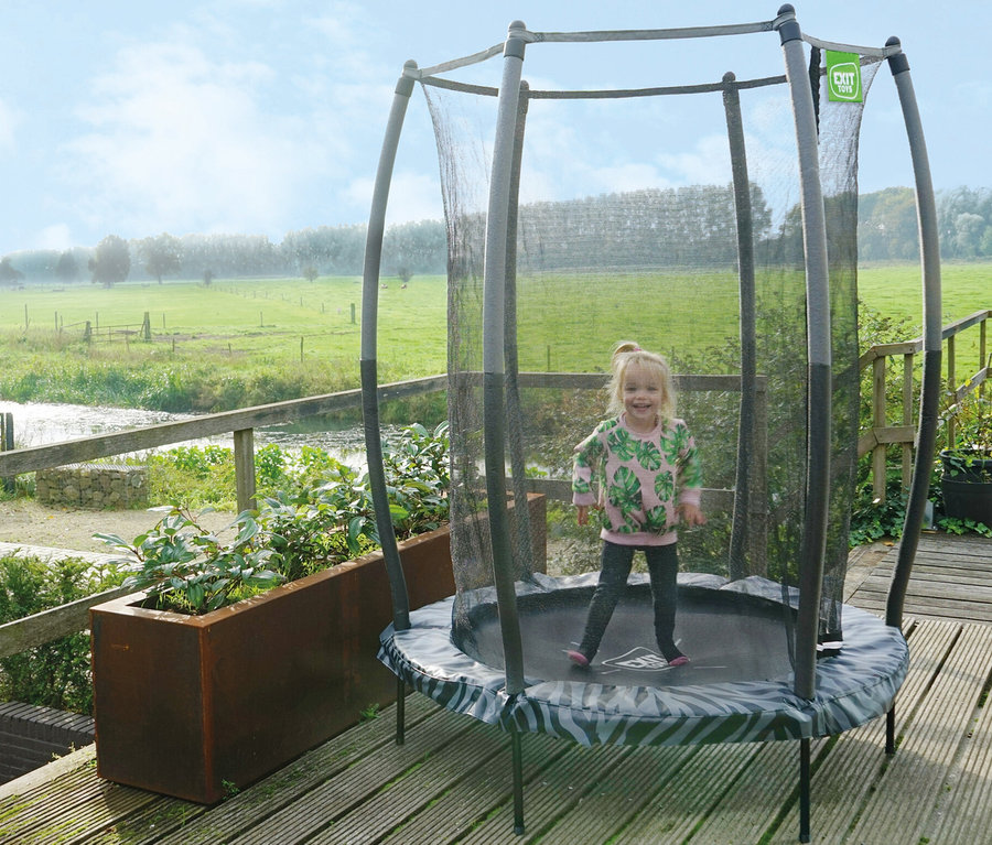 Mädchen springt auf einem EXIT Tiggy Junior Trampolin mit Sicherheitsnetz im Freien auf einer Holzterrasse.