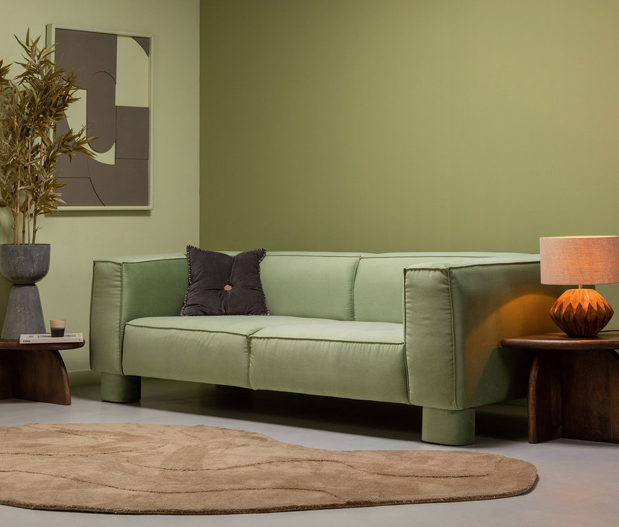 Ein grünes Wohnzimmer mit Sofa, braunem Kissen und braunem, organischem Woood Alya Teppich.