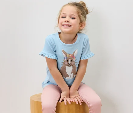 Mädchen sitzt auf einem Holzhocker und trägt ein blaues T-Shirt mit Hasenapplikation.