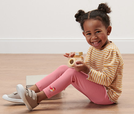 Ein lächelndes Mädchen sitzt auf dem Boden und spielt mit einer Holzkamera. Sie trägt eine geringelte Bluse und 5 Kinder-Leggings mit Alloverprint.