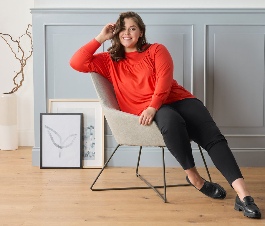 Frau sitzt auf einem Stuhl und trägt eine karierte Stretchhose, Leder-Loafer und ein Blusenshirt mit Raffung, orange-rot.