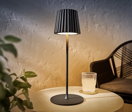 Eine schwarze LED-Akku-Outdoor-Tischleuchte steht auf einem Tisch neben einem Glas mit einem Getränk.