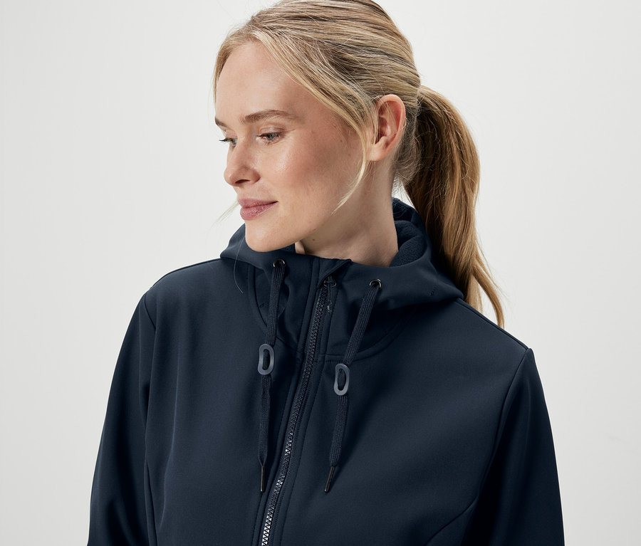 Porträt einer Frau mit blonden, zu einem Pferdeschwanz gebundenen Haaren, die einen dunkelblauen Kapuzenpullover mit Reißverschluss trägt.