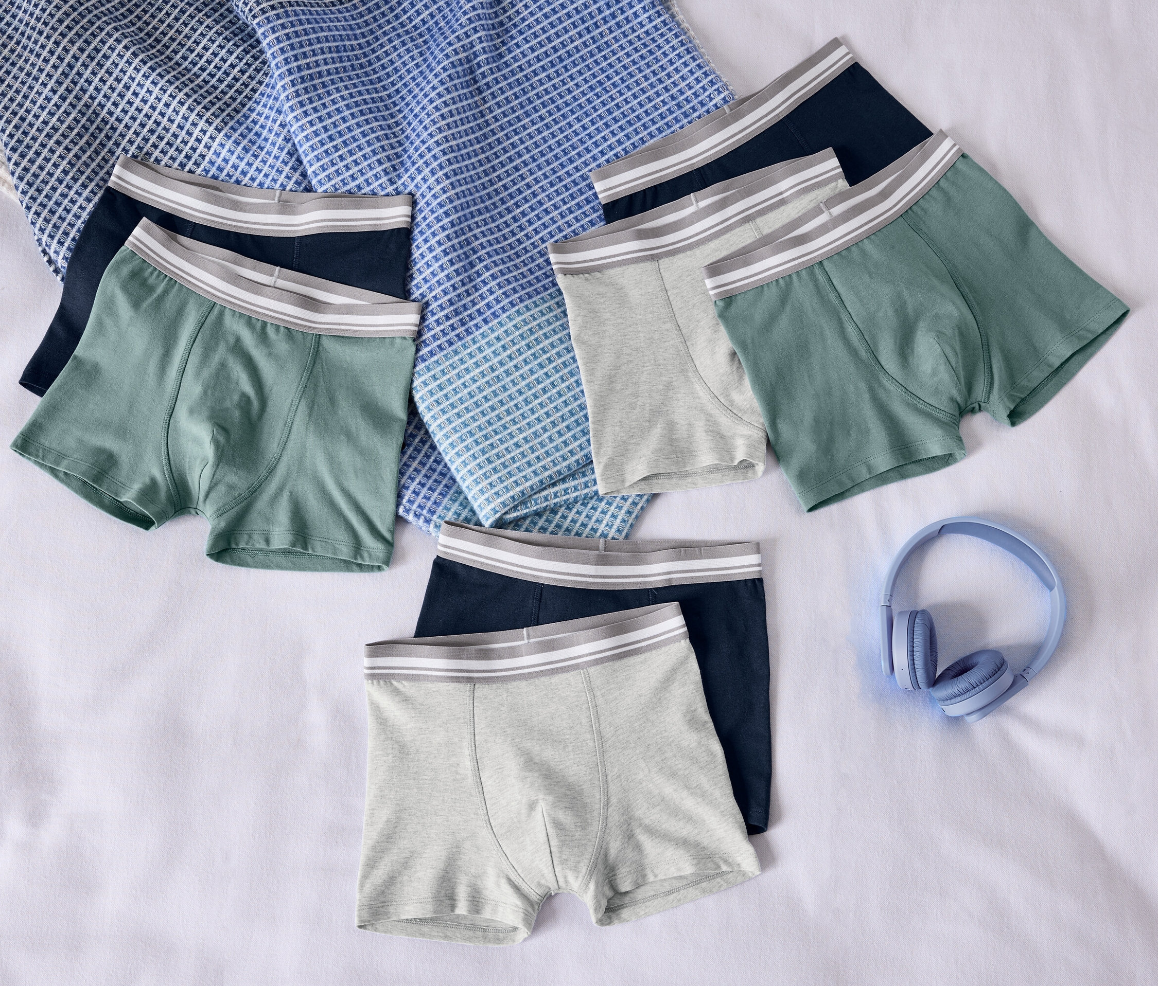 Auf einem weißen Betttuch liegen Herren Boxershorts in Grau, Grün und Dunkelblau sowie hellblaue Kopfhörer.