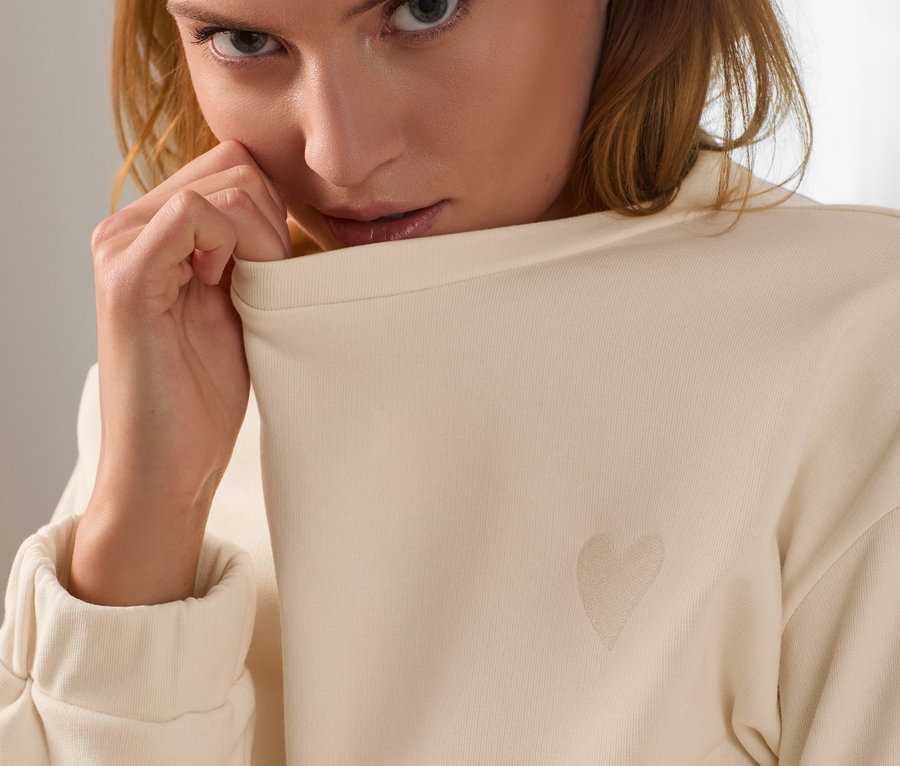 Porträt einer Frau, die den Kragen eines beigen Sweatshirts mit einem dezenten Herz hält.