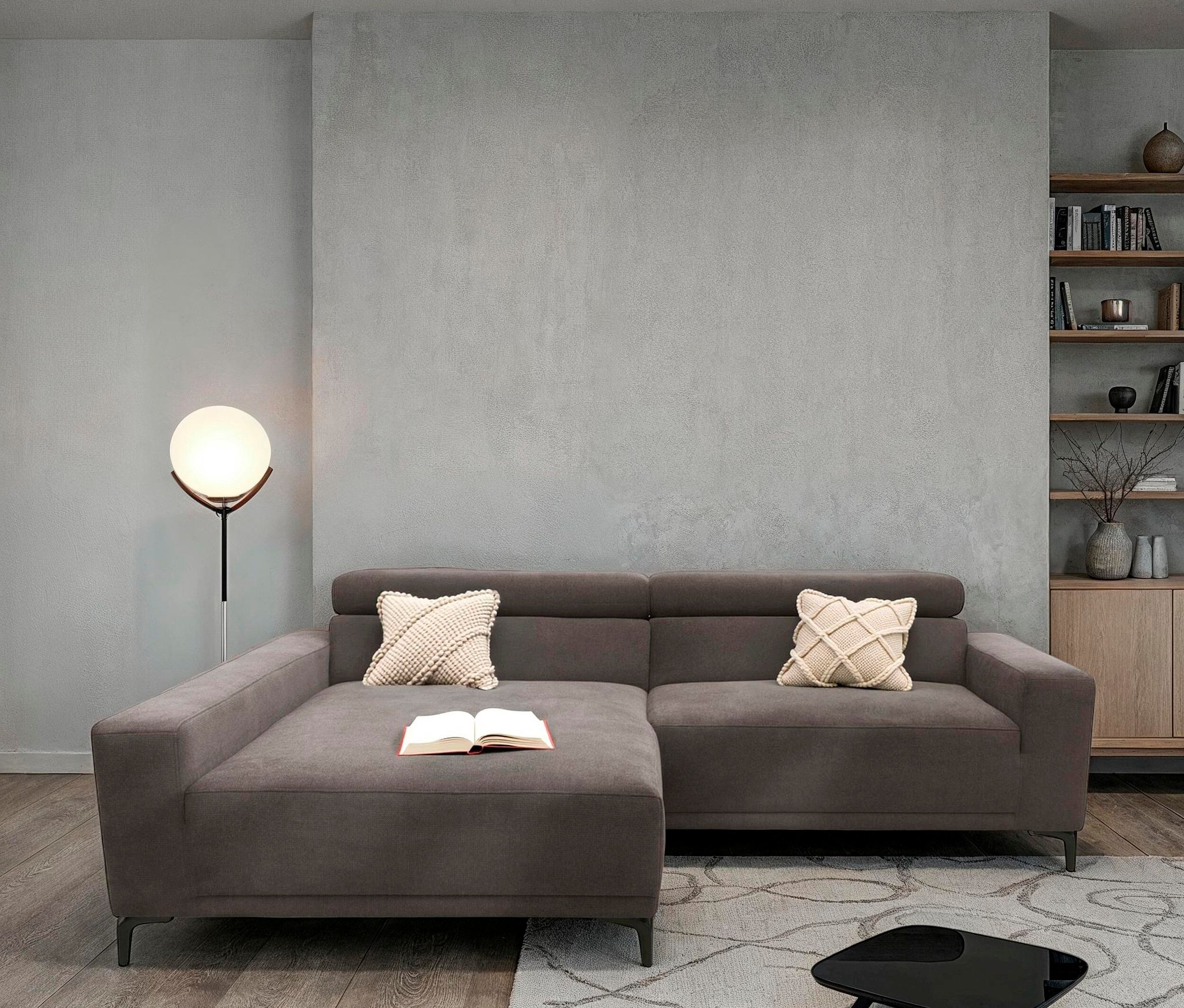 Wohnzimmer mit grauem Ecksofa, Kissen, Buch und Lampe.