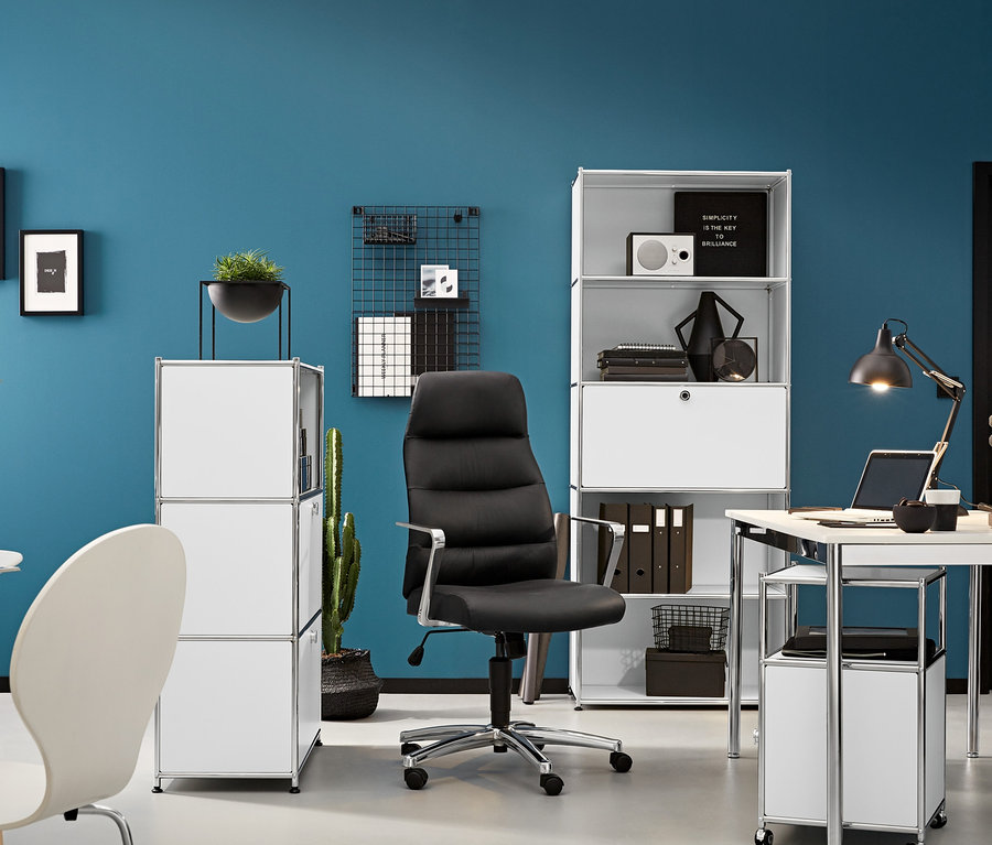 Ein anthrazitfarbener Schreibtischstuhl steht zwischen einem hohen Metall-Regal mit Klappenfach und einem Schreibtisch. Unter dem Schreibtisch steht ein weißer Rollcontainer Metall »CN3« mit Tür.