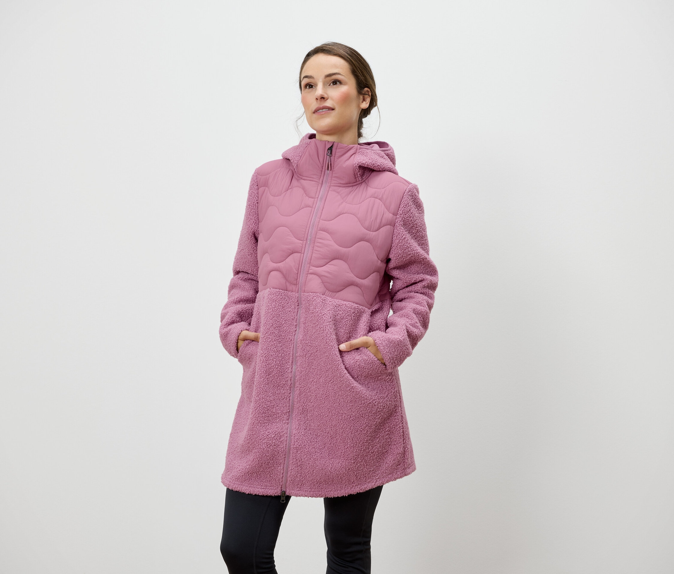 Frau steht mit den Händen in den Taschen eines rosafarbenen Reissverschlussmantels mit Kapuze.