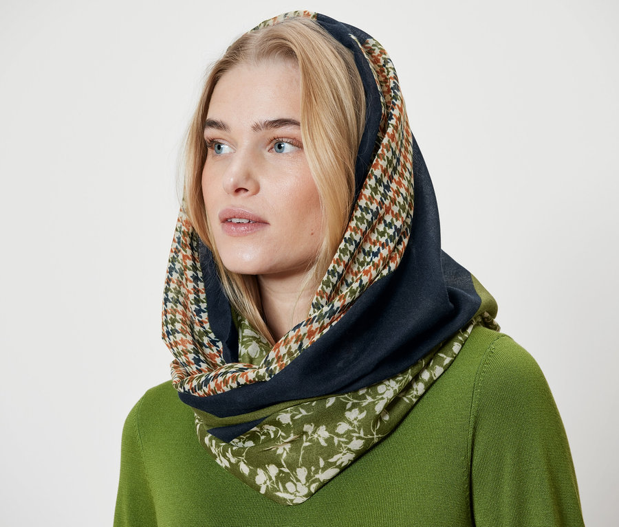 Blonde Frau trägt ein Tuch mit Hahnentritt-, marineblauer und floraler Lage und einen grünen Pullover.
