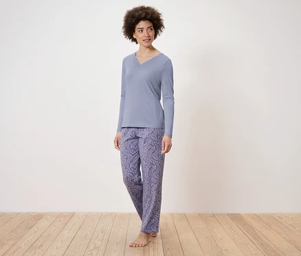 Eine Frau steht und trägt einen blauen, langärmligen Schlafanzug und gemusterte Hosen.