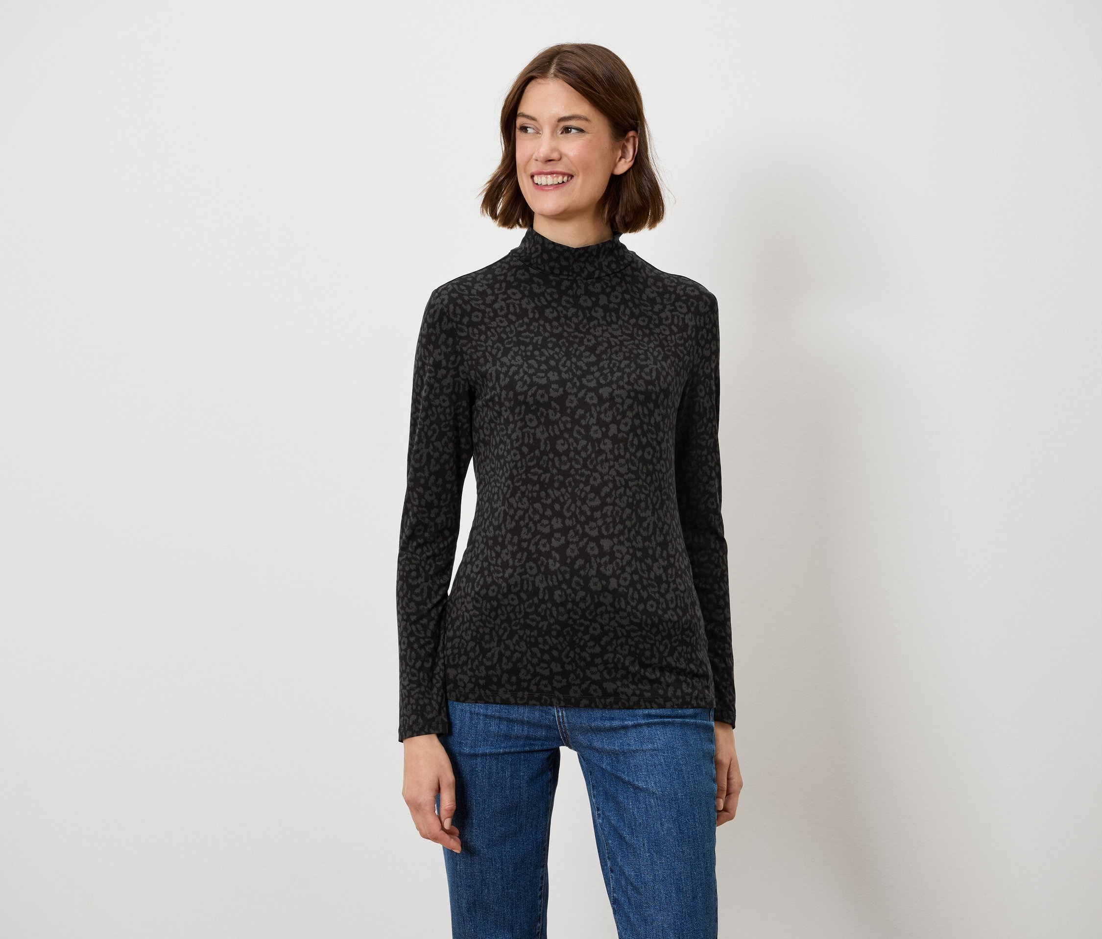 Eine Frau mit braunen Haaren posiert lächelnd in einem schwarzen Langarmshirt und blauen Jeans.