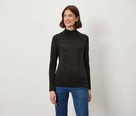 Eine Frau mit braunen Haaren posiert lächelnd in einem schwarzen Langarmshirt und blauen Jeans.