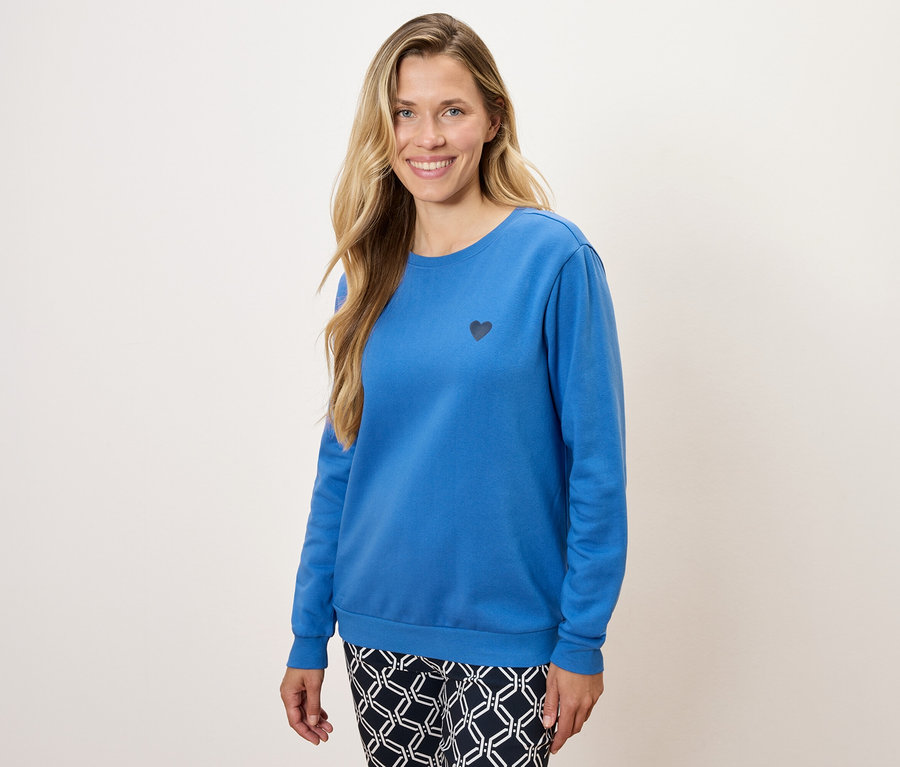 Eine Frau trägt ein blaues Langarmshirt mit einem kleinen schwarzen Herz und gemusterte Hosen.