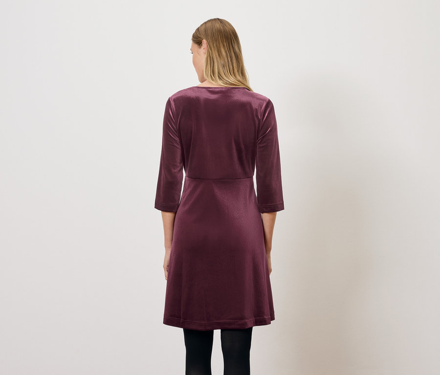 Eine Frau steht mit dem Rücken zum Betrachter und trägt ein weinrotes Samtkleid mit Dreiviertelärmeln.