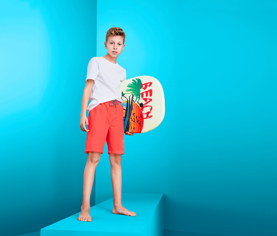 Junge steht auf einer Plattform, hält ein Beach-Board und trägt Boardshorts.